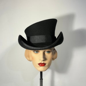 May include: A black felt top hat with a wide brim and a tall crown. The hat is angled to the side and is sitting on a mannequin head.