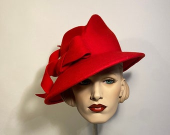 Puede incluir: Un sombrero de fieltro rojo con un gran lazo en un lado. El sombrero está inclinado sobre una cabeza de maniquí.