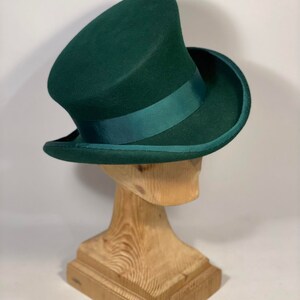 May include: A dark green top hat with a matching ribbon band, displayed on a wooden hat stand. The hat has a tall, cylindrical crown and a wide, curved brim. The hat is a classic style, suitable for formal events.