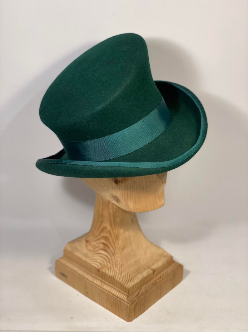 May include: A dark green top hat with a matching ribbon band, displayed on a wooden hat stand. The hat has a tall, cylindrical crown and a wide, curved brim. The hat is a classic style, suitable for formal events.