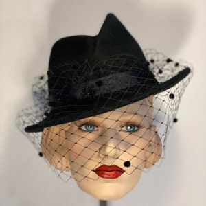 Puede incluir: Un sombrero de fieltro negro con ala ancha y velo negro. El velo tiene peque&ntilde;os puntos negros por todas partes. El sombrero est&aacute; inclinado sobre una cabeza de maniqu&iacute;.