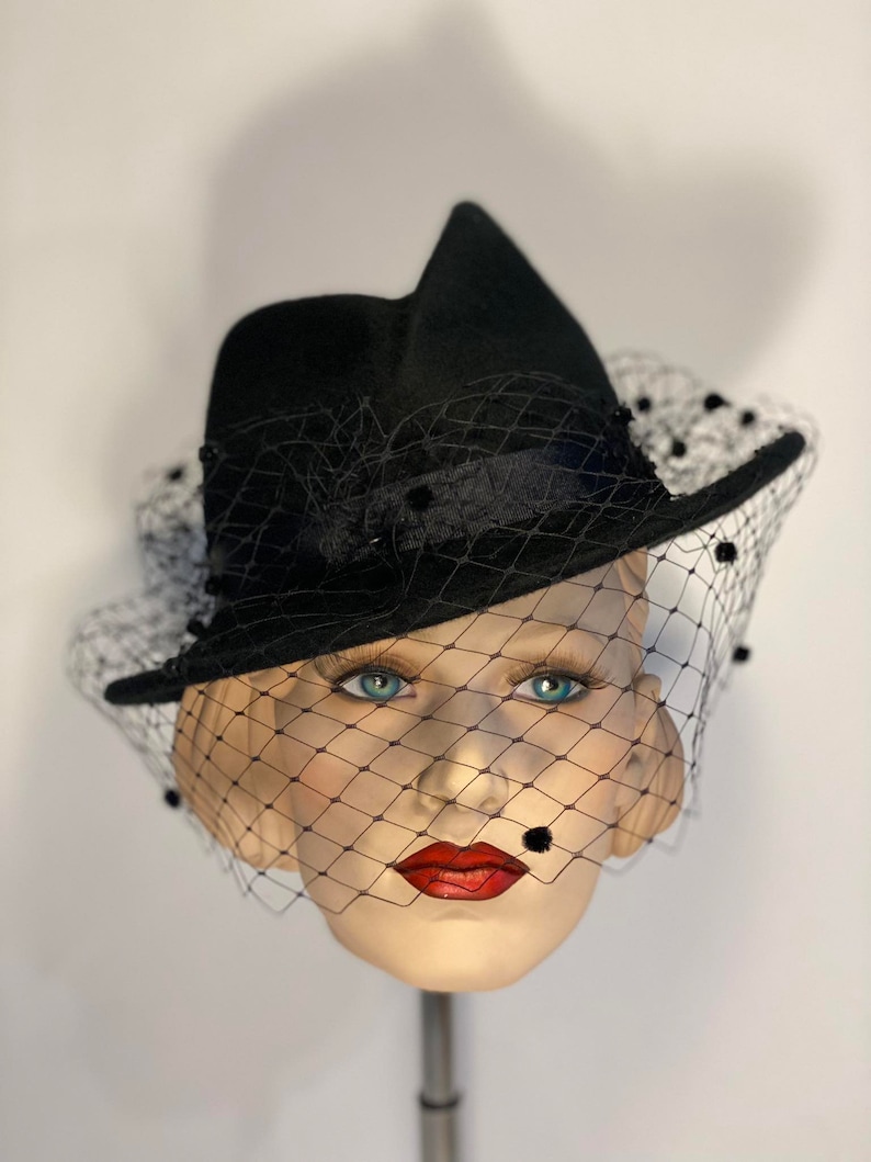 Puede incluir: Un sombrero de fieltro negro con ala ancha y velo negro. El velo tiene peque&ntilde;os puntos negros por todas partes. El sombrero est&aacute; inclinado sobre una cabeza de maniqu&iacute;.