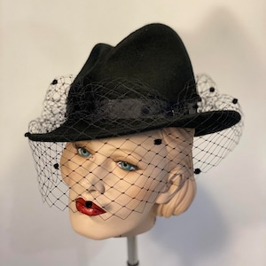 Puede incluir: Un sombrero fedora de fieltro negro con velo negro y lunares negros. El sombrero est&aacute; en una cabeza de maniqu&iacute;.