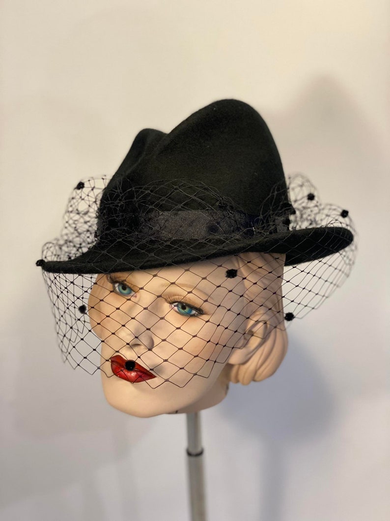 Puede incluir: Un sombrero fedora de fieltro negro con velo negro y lunares negros. El sombrero est&aacute; en una cabeza de maniqu&iacute;.