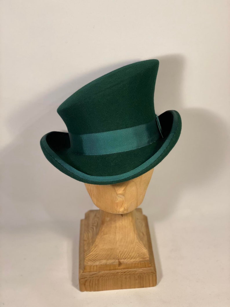 May include: A dark green top hat with a matching green ribbon band. The hat is displayed on a wooden hat stand. The hat has a curved brim and a tall, slightly angled crown. The hat is made of a felt-like material.