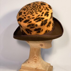May include: A brown top hat with a leopard print crown. The hat is on a wooden hat stand.