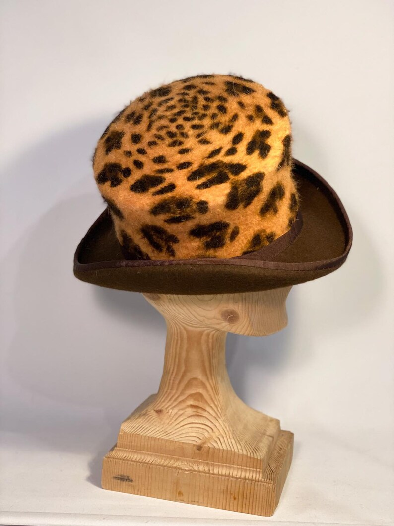May include: A brown top hat with a leopard print crown. The hat is on a wooden hat stand.