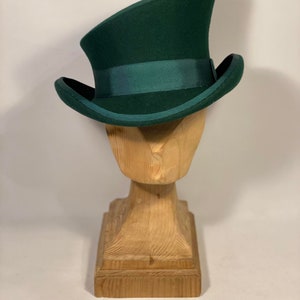 May include: A dark green top hat with a matching green band, displayed on a wooden hat stand. The hat has a curved brim and a tall, cylindrical crown. The wooden stand is carved with a square base and a pedestal.