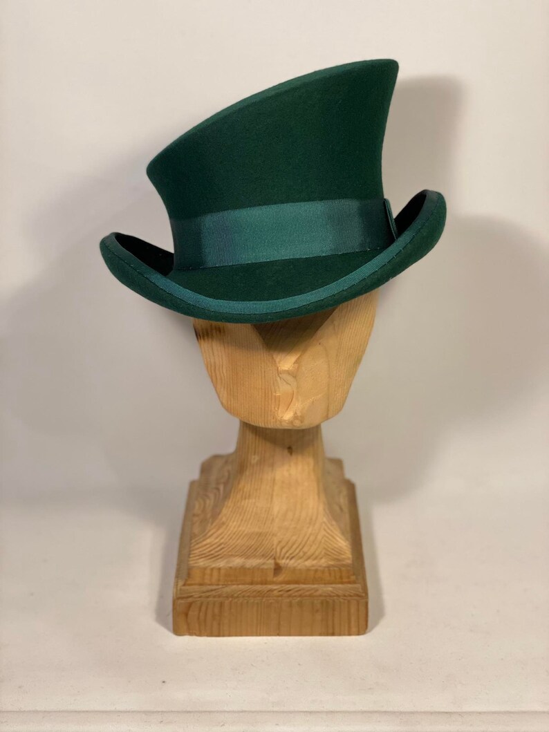 May include: A dark green top hat with a matching green band, displayed on a wooden hat stand. The hat has a curved brim and a tall, cylindrical crown. The wooden stand is carved with a square base and a pedestal.