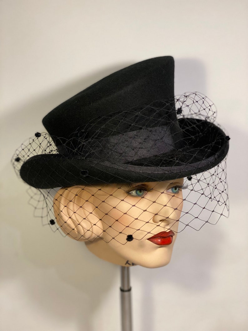 May include: A black top hat with a black veil and black bead accents. The hat is angled on a mannequin head.