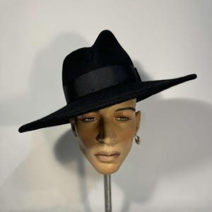Può includere: Un cappello fedora nero a tesa larga e fascia nera. Il cappello è inclinato di lato e si trova su una testa di manichino.