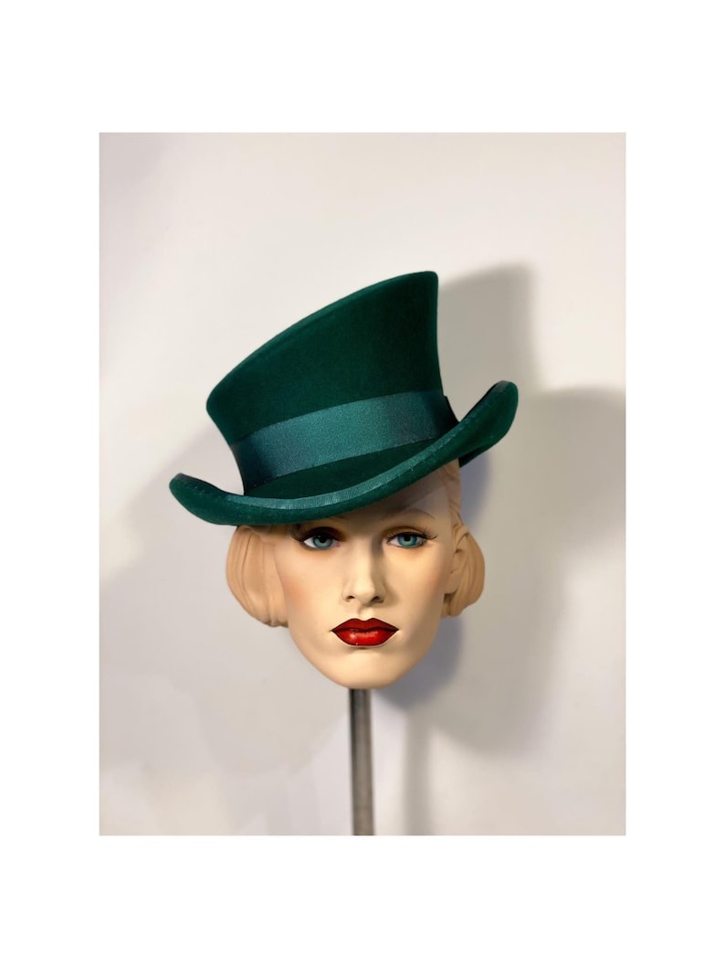 May include: A dark green top hat with a matching ribbon band. The hat is displayed on a mannequin head with blonde hair, blue eyes, and red lipstick. The hat is a classic style.