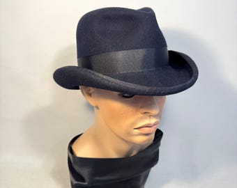 Op de afbeelding: Een donker marineblauwe fedora hoed met een zwarte grosgrain lint band. De hoed wordt getoond op een mannequin hoofd. De hoed heeft een klassieke, licht gebogen rand en een geknepen kroon. De hoed is gemaakt van vilt.