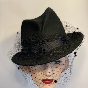 Puede incluir: Un sombrero de fieltro negro con ala ancha y velo negro. El sombrero tiene una forma angular &uacute;nica y est&aacute; decorado con peque&ntilde;as cuentas negras.