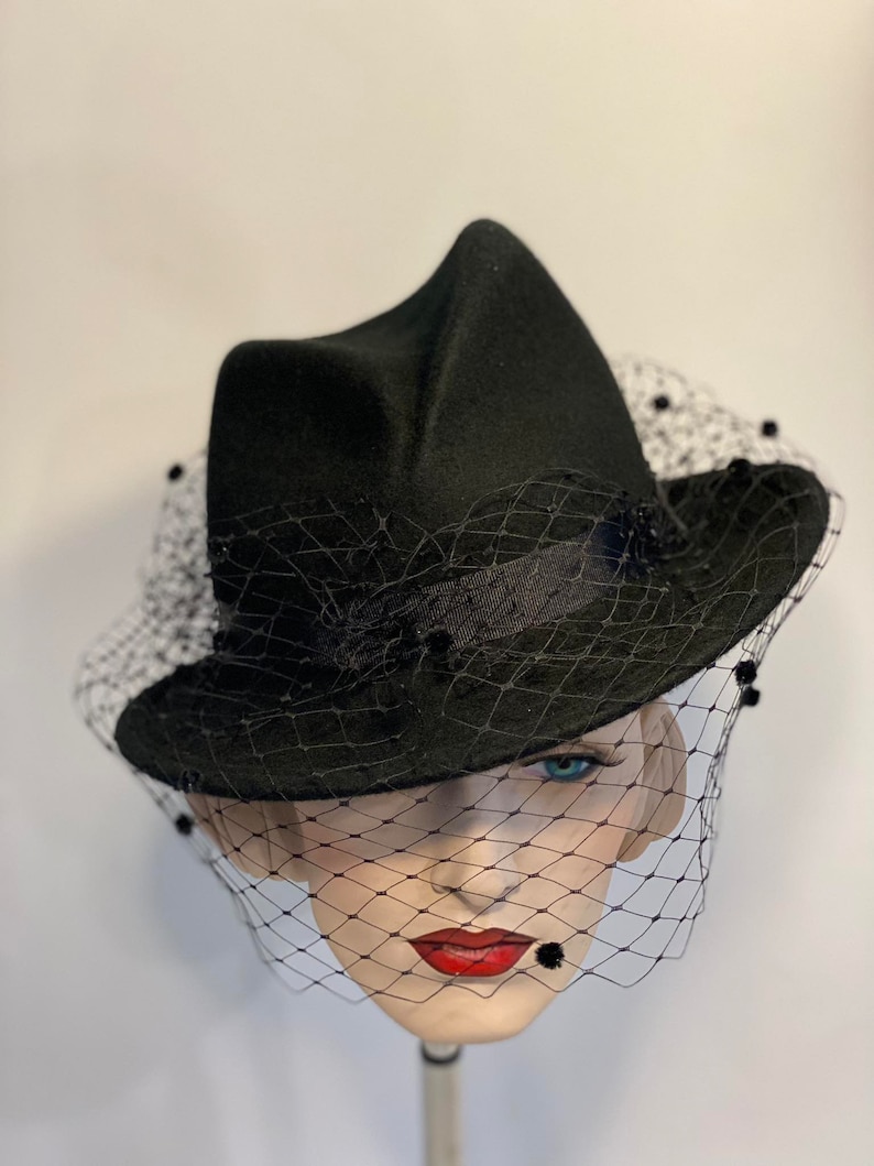 Puede incluir: Un sombrero de fieltro negro con ala ancha y velo negro. El sombrero tiene una forma angular &uacute;nica y est&aacute; decorado con peque&ntilde;as cuentas negras.