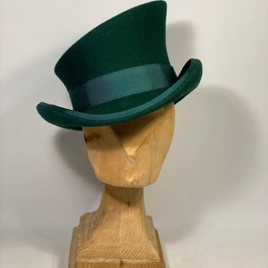 May include: A dark green top hat with a matching ribbon band, displayed on a wooden head form. The hat has a tall, curved crown and a slightly curved brim. The hat is a classic style.