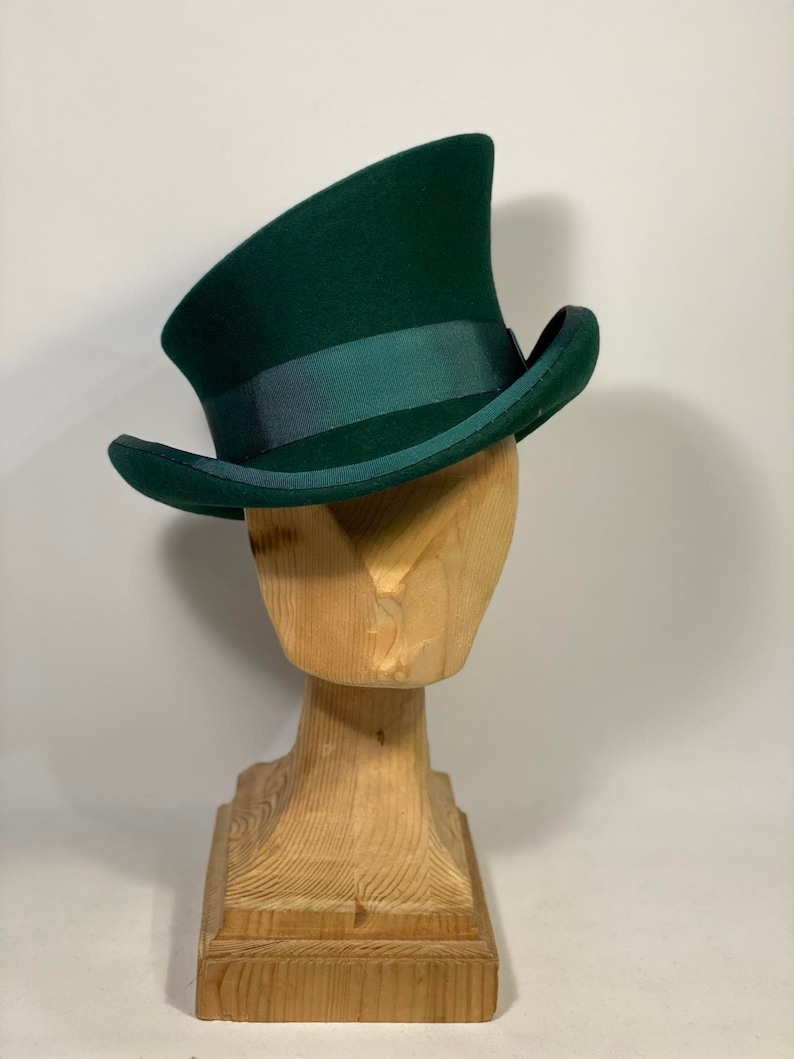 May include: A dark green top hat with a matching ribbon band, displayed on a wooden head form. The hat has a tall, curved crown and a slightly curved brim. The hat is a classic style.