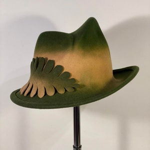 Könnte beinhalten: Ein grüner und brauner Filzhut mit einem einzigartigen Design, das eine Reihe von gewellten Blättern um den Rand herum aufweist. Der Hut wird auf einem schwarzen Ständer präsentiert.