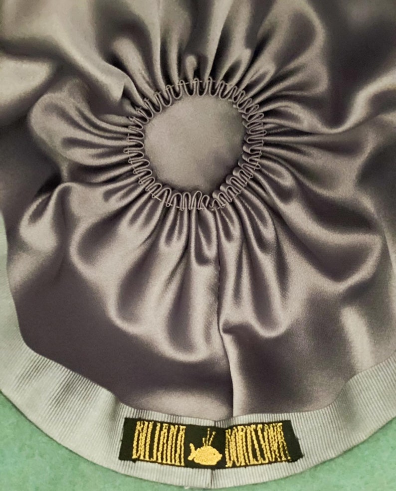 May include: The inside of a teal green felt hat with a gray satin lining and a gray grosgrain ribbon. The ribbon has a gold label with the text "Billini Borse" and a small whale graphic.