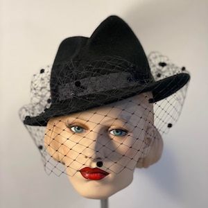 Puede incluir: Un sombrero fedora de fieltro negro con velo negro y adornos de cuentas negras. El sombrero est&aacute; inclinado ligeramente y el velo est&aacute; drapeado sobre la parte delantera del sombrero.