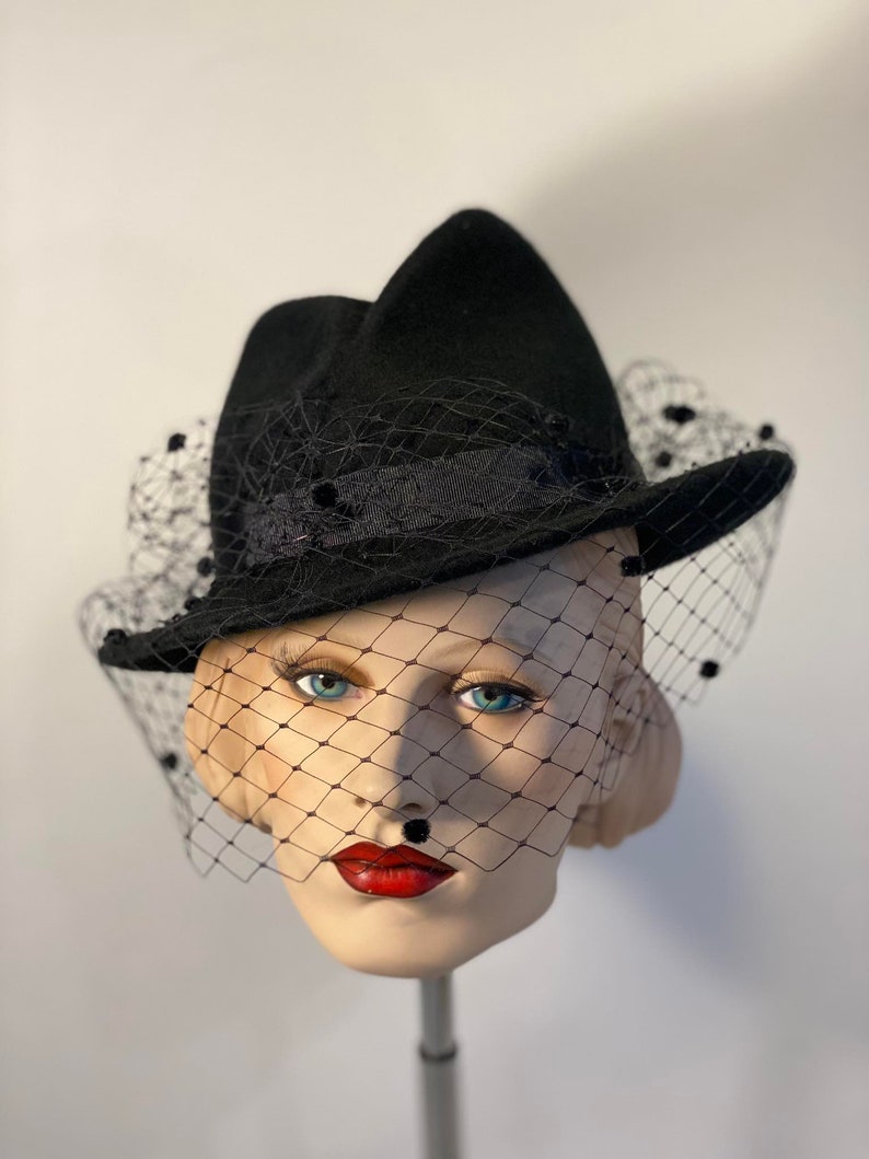 Puede incluir: Un sombrero fedora de fieltro negro con velo negro y adornos de cuentas negras. El sombrero est&aacute; inclinado ligeramente y el velo est&aacute; drapeado sobre la parte delantera del sombrero.