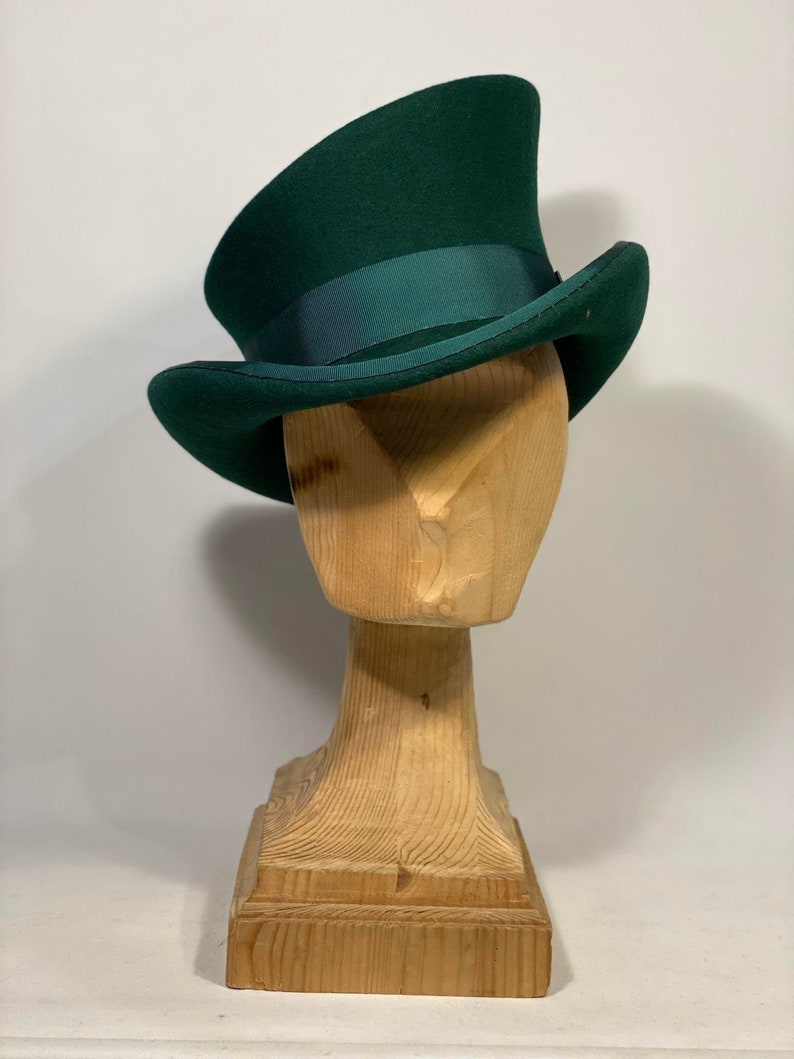 May include: A dark green top hat with a matching ribbon band, displayed on a wooden hat form. The hat has a tall, cylindrical crown and a slightly curved brim. The hat form is made of light-colored wood.