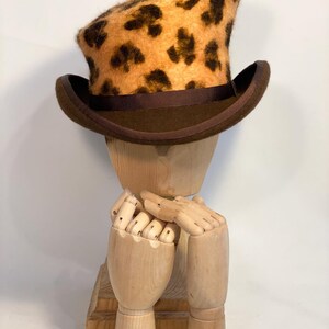 May include: A brown and tan leopard print top hat with a brown brim. The hat is sitting on a wooden mannequin head.