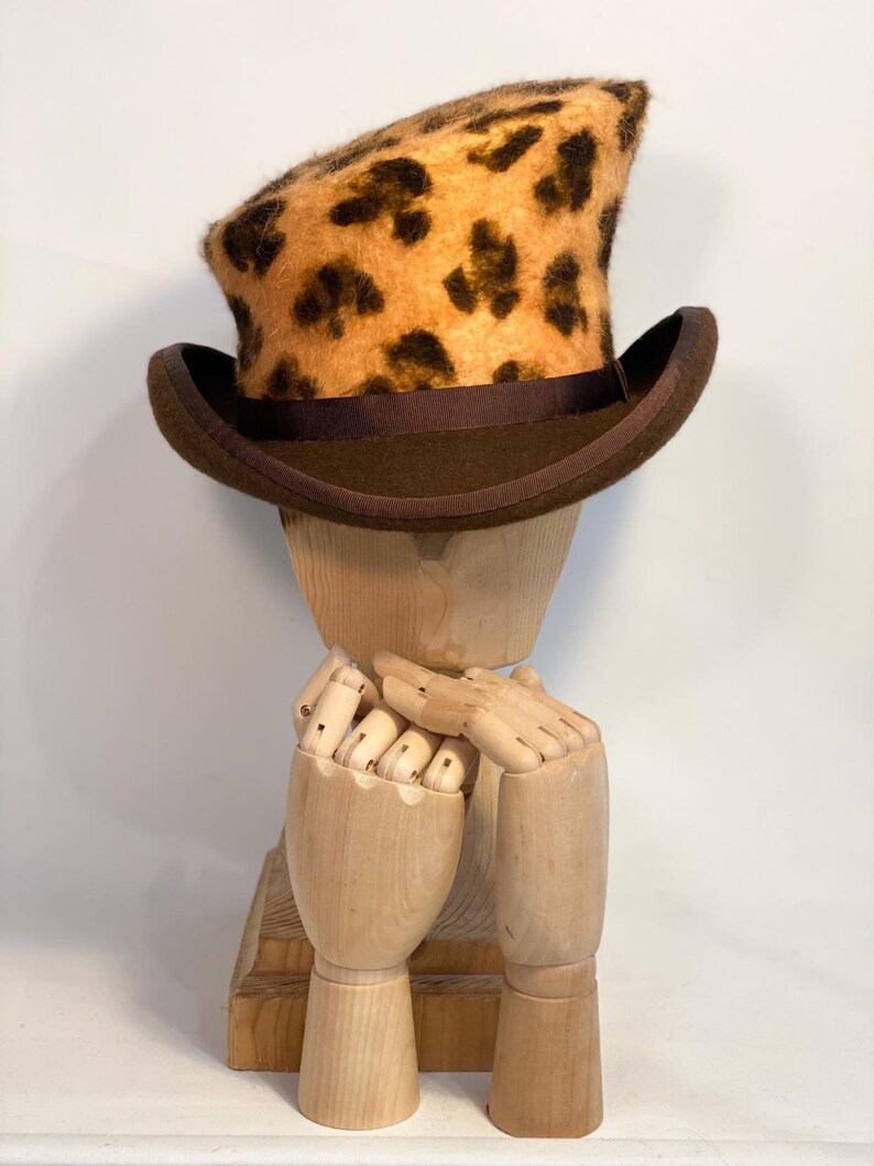 May include: A brown and tan leopard print top hat with a brown brim. The hat is sitting on a wooden mannequin head.