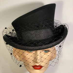 May include: A black top hat with a black veil and black bead accents. The hat is angled to the side and the veil is draped over the front of the hat.