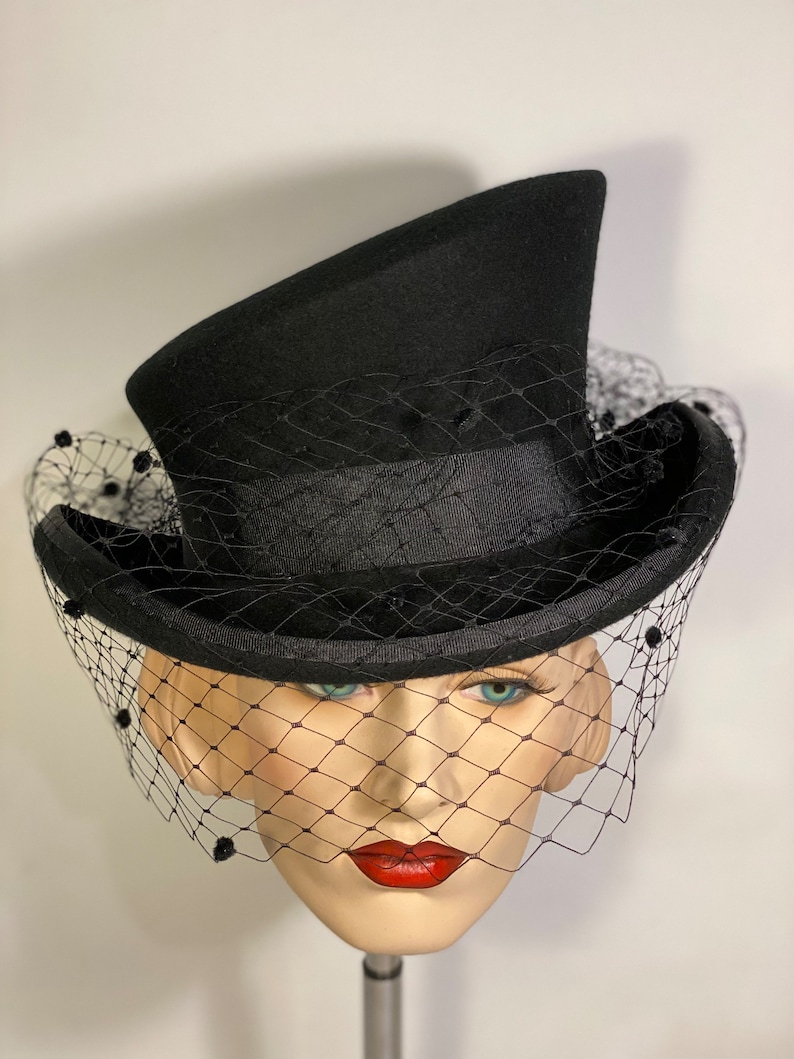 May include: A black top hat with a black veil and black bead accents. The hat is angled to the side and the veil is draped over the front of the hat.