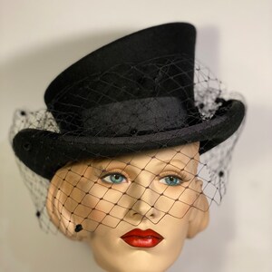 May include: A black top hat with a black veil. The hat is sitting on a mannequin head.