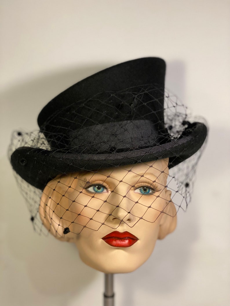 May include: A black top hat with a black veil. The hat is sitting on a mannequin head.