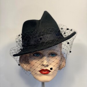 Puede incluir: Un sombrero fedora de fieltro negro con velo negro y adornos de cuentas negras. El sombrero est&aacute; inclinado sobre una cabeza de maniqu&iacute;.