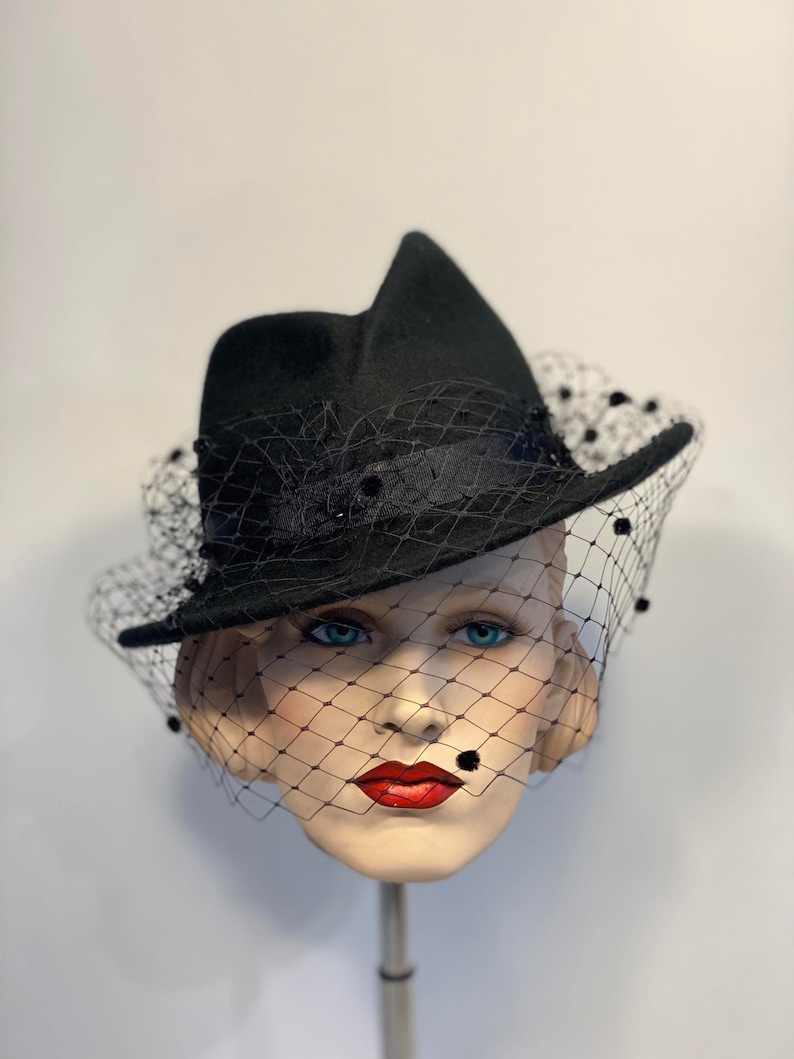 Puede incluir: Un sombrero fedora de fieltro negro con velo negro y adornos de cuentas negras. El sombrero est&aacute; inclinado sobre una cabeza de maniqu&iacute;.