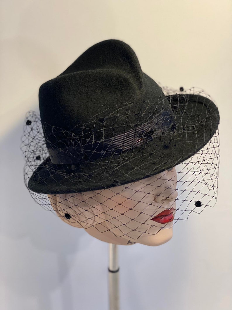 Puede incluir: Un sombrero de fieltro negro con ala ancha y velo negro. El sombrero tiene una forma &uacute;nica y est&aacute; decorado con peque&ntilde;os puntos negros.