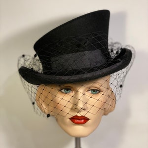 May include: A black top hat with a black veil. The hat is angled to the side and the veil is covering the face of a mannequin.