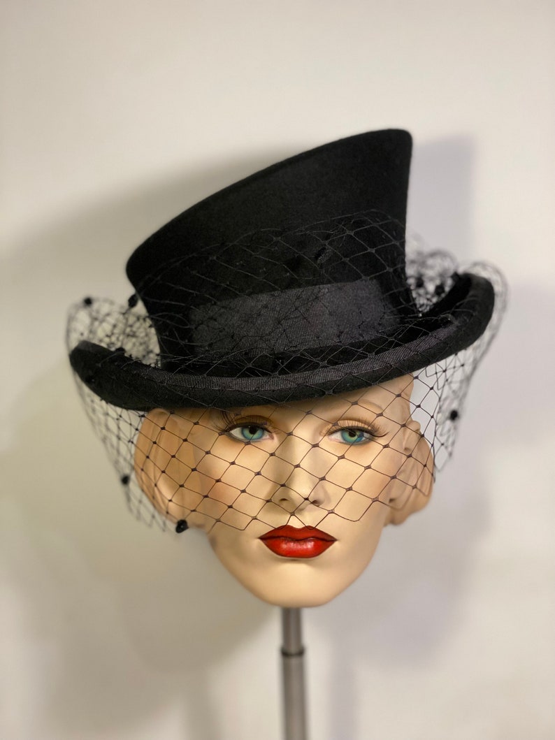 May include: A black top hat with a black veil. The hat is angled to the side and the veil is covering the face of a mannequin.
