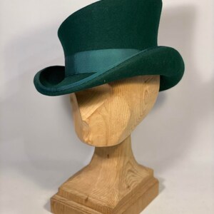May include: A dark green top hat with a matching ribbon band. The hat is displayed on a wooden head form. The hat has a curved brim and a tall, cylindrical crown. The hat is a classic style.