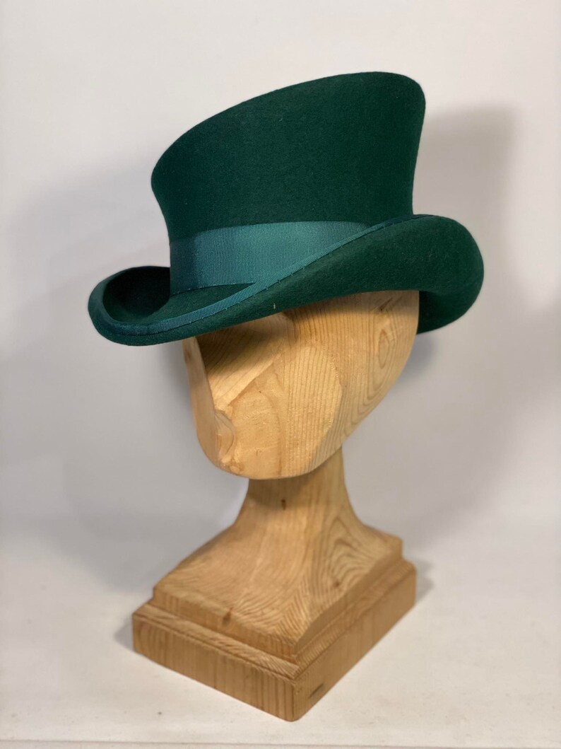 May include: A dark green top hat with a matching ribbon band. The hat is displayed on a wooden head form. The hat has a curved brim and a tall, cylindrical crown. The hat is a classic style.