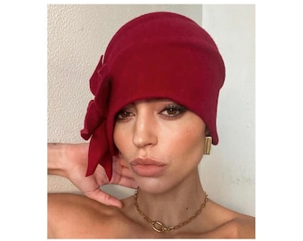 May include: A woman wearing a red felt hat with a large flower detail. The hat is angled on her head and covers part of her forehead. She is wearing a gold chain necklace and gold earrings.