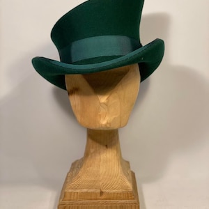 May include: A dark green top hat with a matching ribbon band, displayed on a wooden hat stand. The hat has a classic tall crown and a slightly curved brim. The wooden stand is carved in a stylized bust shape.
