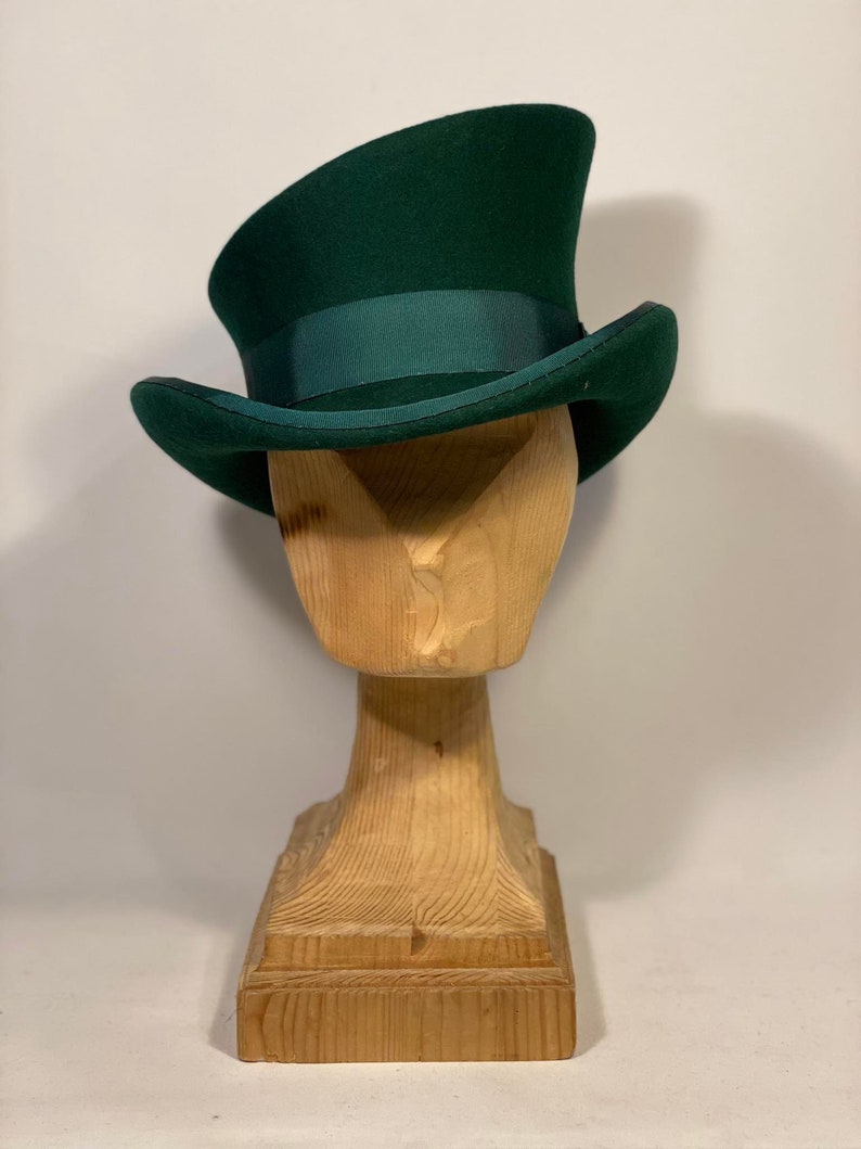 May include: A dark green top hat with a matching ribbon band, displayed on a wooden hat stand. The hat has a classic tall crown and a slightly curved brim. The wooden stand is carved in a stylized bust shape.