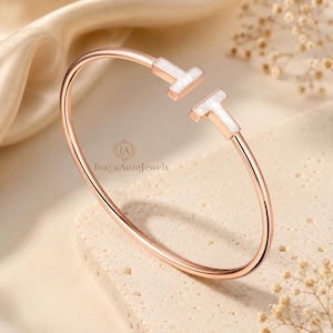Könnte beinhalten: Ein offener Armreif in Roségold-Optik mit zwei T-förmigen Endstücken, die mit weißem Material eingelegt sind. Der Armreif zeigt mittig den Text InayaAuraJewels. Das Design besteht aus einem dünnen, polierten Metallband in kreisförmiger Form.