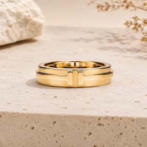 Könnte beinhalten: Ein polierter Goldring mit einem zentralen, horizontalen T-förmigen Motiv, das die beiden Seiten des Bandes verbindet. Der Ring hat eine glatte, reflektierende Metalloberfläche und eine gleichmäßige Breite, was ein klares, geometrisches Design ergibt.