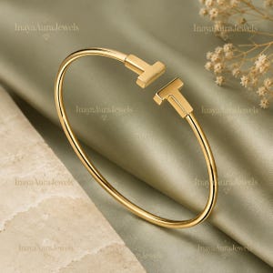 Può includere: Un bracciale aperto color oro caratterizzato da due terminali a forma di T rivolti l'uno verso l'altro nell'apertura centrale. Il cinturino in metallo ha una finitura liscia e lucida e una forma circolare, creando un design pulito e semplice.