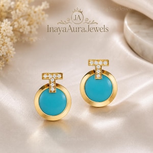 Puede incluir: Un par de pendientes de oro con una piedra turquesa circular engastada en un marco de oro pulido. Una barra en forma de T adornada con pequeños diamantes pavé transparentes se sitúa sobre cada círculo. La marca InayaAuraJewels es visible.