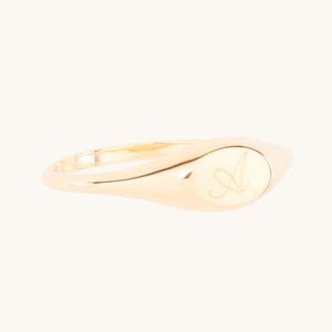 Può includere: Un anello con sigillo d'oro con una fascia liscia e lucida e una faccia ovale. La faccia dell'anello presenta la lettera "A" in corsivo in un colore oro più chiaro. L'anello ha un design classico ed elegante.