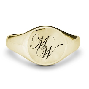 May include: A gold signet ring with a polished finish. The ring features an oval face with the engraved initials "MW" in a delicate script. A classic piece of jewellery.