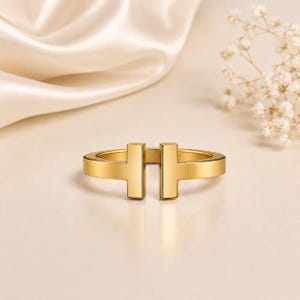 May include: A gold-toned open ring featuring a T-shaped design on each end. The band has a smooth, polished finish with a gap between the two T-shaped terminals, creating a continuous, sleek, and minimalist circular structure for the finger.