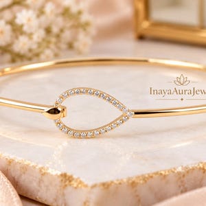 May include: A gold bangle bracelet featuring a central teardrop-shaped motif encrusted with small clear stones. The bracelet has a smooth, polished finish and includes the text InayaAuraJewels printed in a small font near the teardrop design.